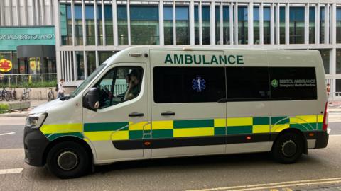 A white ambulance van captured from the left side, with hi-vis yellow and green decals along the bodywork and the word 'Ambulance' at the top on capitals. In the background you can see the Bristol Royal Infirmary building with large glass windows. 
