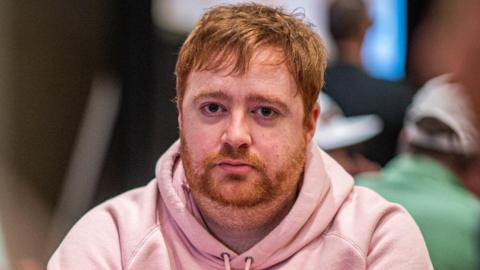 A man with ginger hair and a beard in a pink hooded top stares into the camera