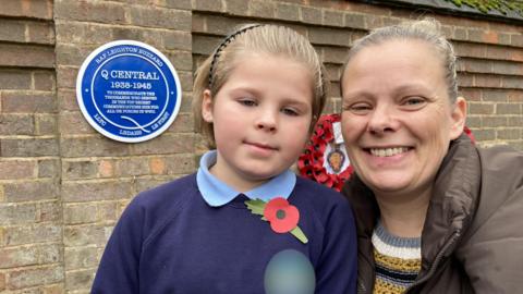 A child wearing a poppy is photographed in front of a blue plaque with a woman smiling next to him
