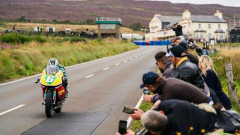 Michael Dunlop on a classic machine riding with fans looking on from the hedgerow and the Creg Ny Baa public house in the background.
