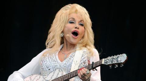 Dolly Parton wearing a sheer white blouse with balloon sleeves and white trousers holding a white and silver acoustic guitar on stage singing into a microphone attached to her head.