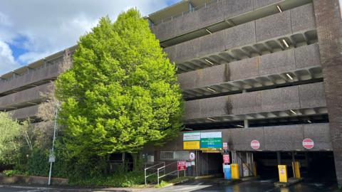 The image shows the exterior of a multi-storey car park. A large green tree is in front of the concrete car park.