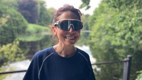 A smiling woman wearing dark sunglasses and a navy T-shirt. In the backdrop are railings with a river running under, surrounded by greenery.