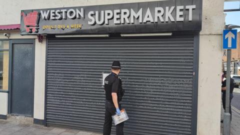 A police officer wearing a black uniform, hat and blue latex gloves. He is standing outside the Weston Supermarket, attaching a closure order to the black corrugated shutters.