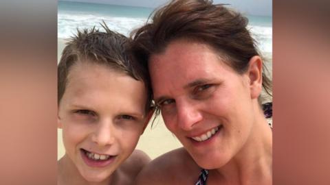 A mum and her young teenage son smile for a selfie as they stand on a beach abroad with the ocean behind them