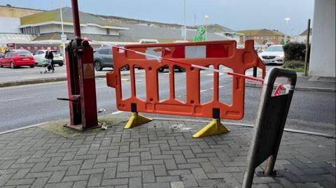 A red plastic barrier in place at the edge of a car park with shoppers and cars in the background. 