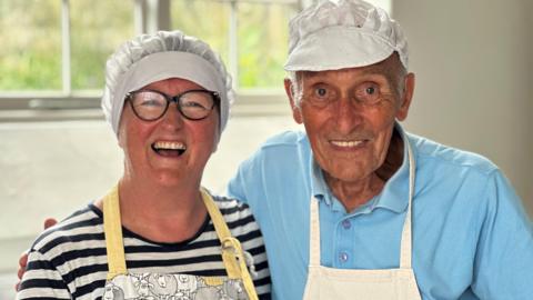 Fenella Costain, who is wearing a black and white striped top and glasses, standing next to Peter Girdler who is wearing a light blue t-shirt and has his arm around her. Both are wearing white chefs hats and aprons.