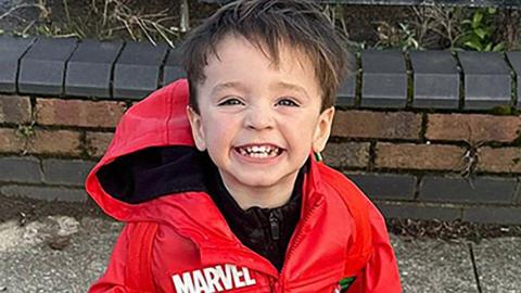 Jude Gerrard, a young boy with brown hair, grins as he is being photographed. He is wearing a red waterproof coat with Marvel branding.