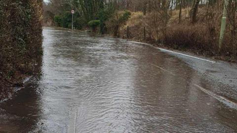 A road with brown hedges and trees on either side. There is a 40 mile per hour road sign on the right hand side. There is flood water covering the entire width of the road, stretching around a slight bend.