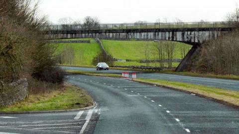 Underbarrow Road Bridge over the A591. A single car drives on the other side of the road. There is a short patch of grass in between the double carriageway. There is a small junction to the left.