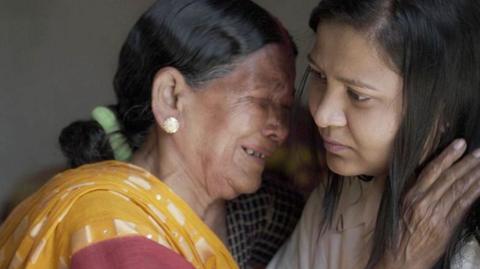 Siro, an elderly midwife wearing a yellow sari, sobs as she clings on to Monica - a woman with long dark hair in her late 20s