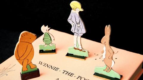 A Winnie-The-Pooh script sits on a table, with little cut outs of Pooh, Piglet, Rabbit and Christopher Robin sitting on top of the script.