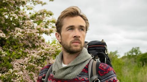 A close-up shot of James, a young bearded gentleman, standing in a field full of green shrubbery and pink flowers. He is wearing a fleece with red, blues and grey on it, and is carrying a rucksack.