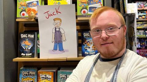 Jack Dillon smiles at the camera next to an illustration of himself from the book. He has short ginger hair and a beard, blue eyes and is wearing glasses, a white shirt and a blue apron. 