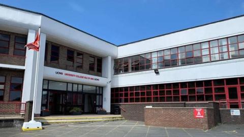 The entrance to the UCM building, which is build of brown bricks and features while concrete cladding and pillars, The entrance is glass-fronted and has UCM University College Isle of Man written in black and red above it.