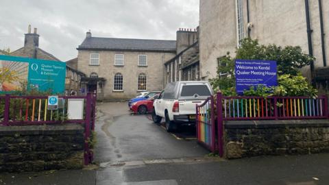 A set of gates painted in rainbow colours are open in the foreground. Beyond them there is a car park with various vehicles in it. In the background there is a grey building with arched windows on the ground floor and square ones on the first. Either side of the entrance gates are two signs, one green and one blue. The former reads Quaker Tapestry Museum and Exhibition while the latter, on the right, says Welcome to Kendal Quaker Meeting House.