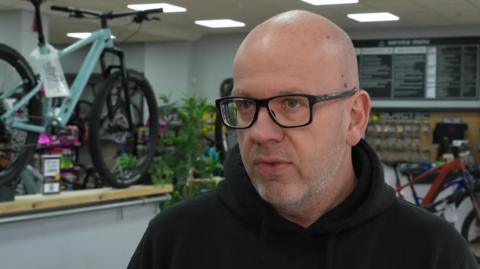 Grant MacIver. He is bald and wears dark-rimmed glasses. He is standing in his bike shop.