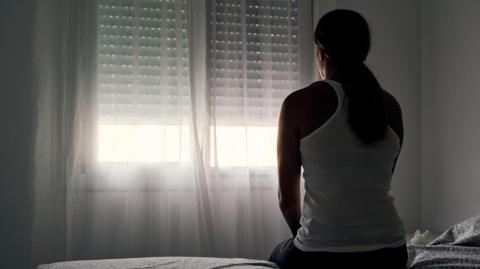 A woman with long brown hair in a ponytail is sat on the edge of the bed facing a white wall and window.