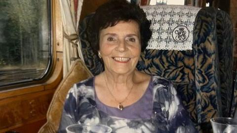 Dorothy Chiles is pictured smiling sat down in a chair. She has short curly brown hair and is wearing a purple flower long sleeved top.
