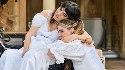 Two women in white wedding dresses hug each other, they are outside on a stone patio