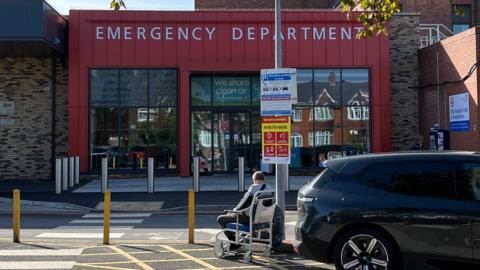 The exterior of Scunthorpe A&E department has brown brickwork and red cladding. A large sign reads Emergency Department. A black car is in the foreground and a patient in a wheelchair is facing the building.