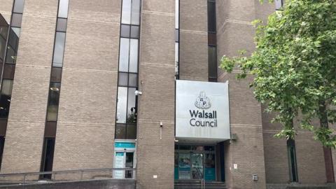 A large brick building with lots of glass panels and a sign reading "WalSALL council" with a tree to the right