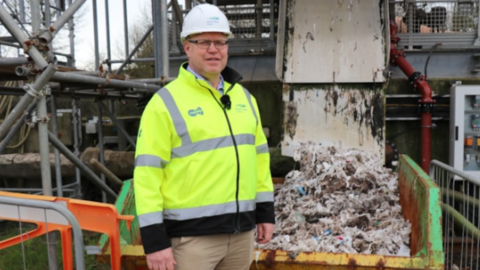 Mr Harris is wearing a hi-vis jacket and a white hard hat. He is stood in front of machinery which appears to have removed waste from drains. 