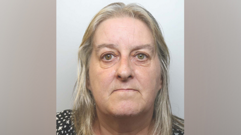 A woman with grey blonde hair stares at the camera in a police mugshot