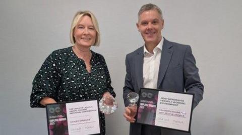 Hayley Douglas wearing a green dress with white patterns holding a certificate and a glass award standing next to Matthew Warren who is wearing a white shirt and a grey jacket also holding the certificate and award.