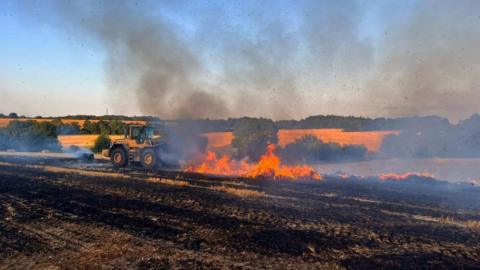 Large fire on a farm, there is a yellow tractor and lots of smoke in the photo