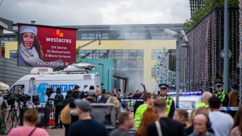 Crowds of people, some dressed as police officers and television journalists, stand outside West Herts College which has been decorated with Christmas lights and a large sign advertising a fictional shopping destination called Westacres.