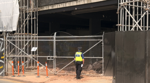 A police officer with their back to the camera close to a road is standing near silver gates and there are vertical silver beams behind the gates and to the right of them on the photo. The next storey is partly visible at the top of the image.