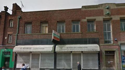 A two-storey red brick art deco style terrace building with an aged white awning that says E. Marsh, with a To Let sign above it. People whose faces have been blurred walk past.