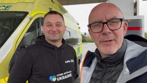 A bald man wearing glasses is taking a selfie with a man wearing a black hoodie with the words Stand With Ukraine written across the chest. The pair are stood in front of an ambulance on what appears to be a petrol forecourt.