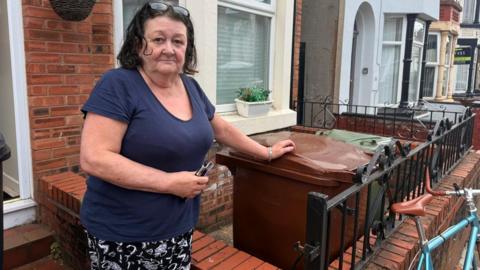 Janette Smith is standing in front of her terraced property, with a brown and a green wheelie bin behind a small brick wall at the front. She has bobbed brown hair and is wearing a blue top and black and white patterned trousers.