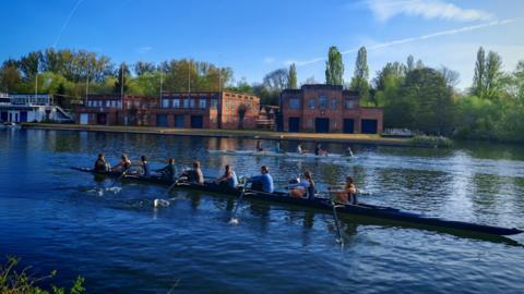 Two rowing boats - a four and an eight rowing along calm water with buildings on the river bank
