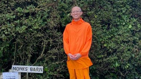 Karucha Sripramote smiling outdoors in front of a dense green hedgerow. His is wearing glasses and a vivid orange outfit comprising a lightweight long-sleeved top and loose knee-length shorts. He is standing with his hands clasped in front of him. A rustic wooden sign reading 'MONKS BARN' is visible to the lower left.