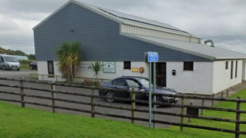 A picture of the community centre. It is a large white and grey building with cars parked around it and a large field next to it. 