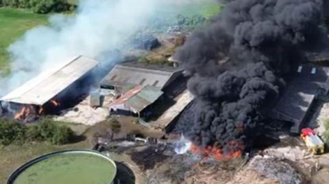 A drone still of a number of farm buildings from above - two are on fire and there are large plumes of smoke, one of which is very dark.