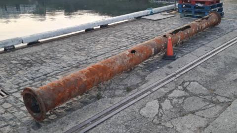 A rusted pipe with holes at the far end. It is lying on the floor at Guernsey's St Peter Port Harbour and the sea can be seen in the top left corner. 
