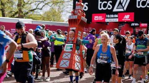 Tony Howarth dressed as The Blackpool Tower finishing the London Marathon. He is surrounded by runners smiling, hugging friends and checking their watches. Tony has his arms outstretched through the sides of his orange coloured costume, and his head popping through a window about half way through the tower. It reaches his knees.