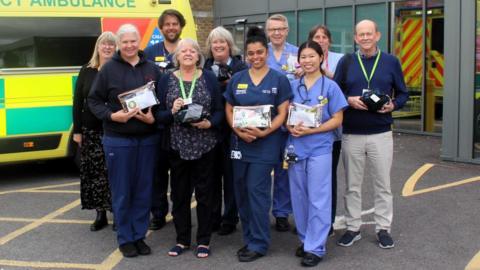 About ten hospital staff - a mixture of men and women, standing next to an ambulance. Some are wearing blue scrubs and lanyards. They are holding clear bags containing toiletries