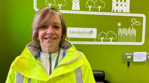A woman wearing a high visibility jacket smiles at the camera. She is standing in front of a green wall with white illustrations and the words "Lincolnshire County Council".