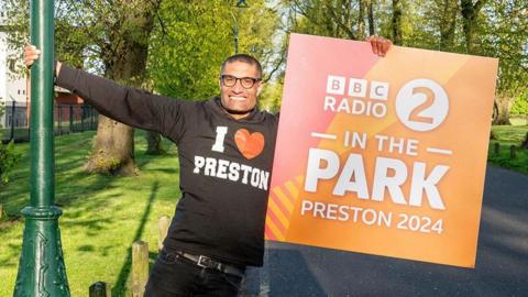 Radio 2's Richie Anderson holding a sign which announces BBC Radio 2's flagship live music festival In The Park will take place in Moor Park, Preston