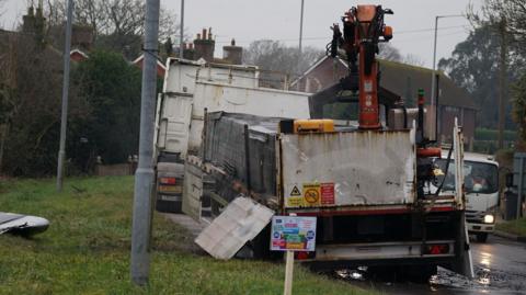 A heavy goods vehicle with a load of what appears to be pallets on the side of a road with a grassy area to the left of it and a house on the road ahead