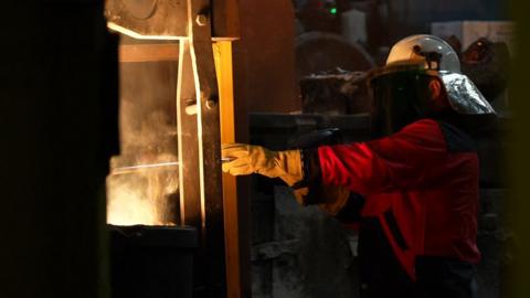 A worker dressed in red protective overalls, brown gloves and a white helmet with a mask stands side on to a piece of machinery from which smoke is visible. His figure is partly in shadow