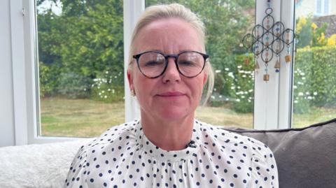 A woman is wearing glasses and is sat inside a house on a sofa. Behind her is a garden. She is wearing a polkadot top and glasses.