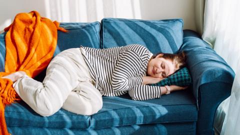 A woman lays on a blue sofa napping, looking content. There is a bright orange blanket thrown over the left hand side of the sofa.
