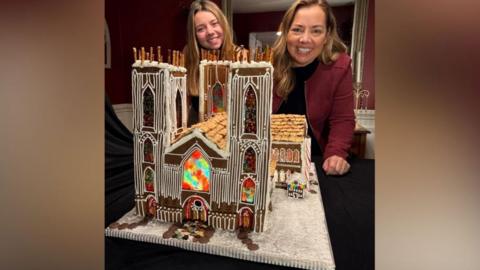 The photo shows a gingerbread cathedral displayed on a table. Tall twin towers are decorated with vertical white icing lines that mimic Gothic stonework. The model has a large central façade window in reds, greens, and yellows and a shingled roof created from cereal pieces.
The gingerbread cathedral sits on a Royal icing base. Two women each with long blonde hair stand behind the creation.