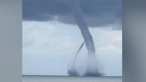 Two large dark grey waterspouts reach up into a dark cloud above the sea off the Italian coastline.